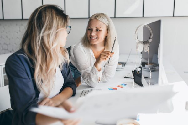 Smiling short-haired blonde girl talking with colleague in office while playing with pencil. Indoor portrait of female web-designer looking at cheerful woman in white shirt.